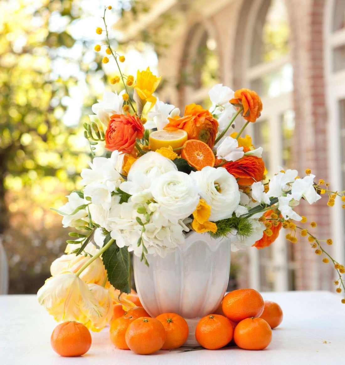 Spring flower arrangement of white and orange blooms in a fluted white vase, with oranges at its base