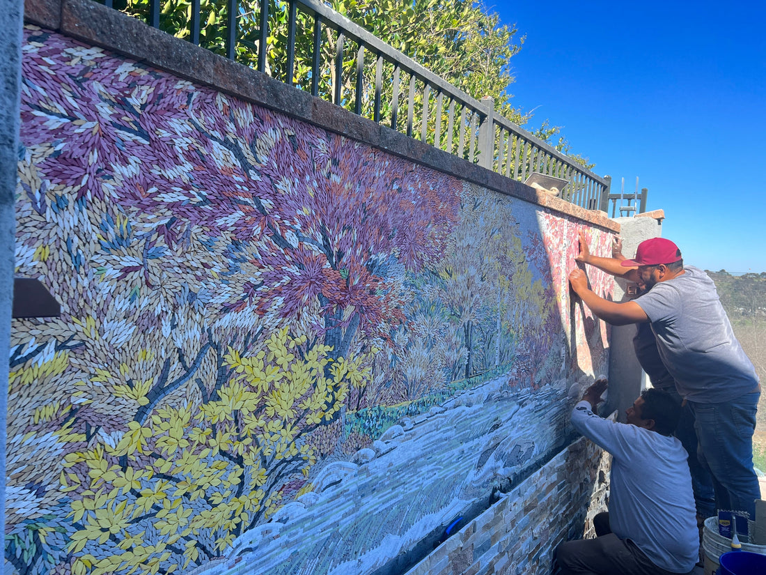 Two people installing a long colourful mosaic mural along a garden wall