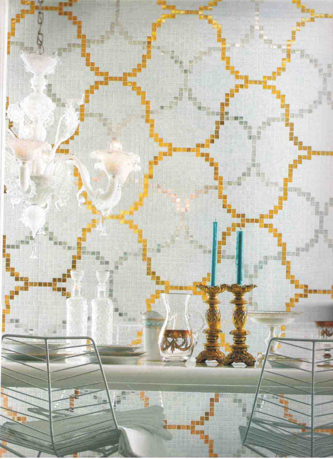 Dining room wall tiled in white mosaic with a gold ogee trellis pattern, behind a glass table and chandelier
